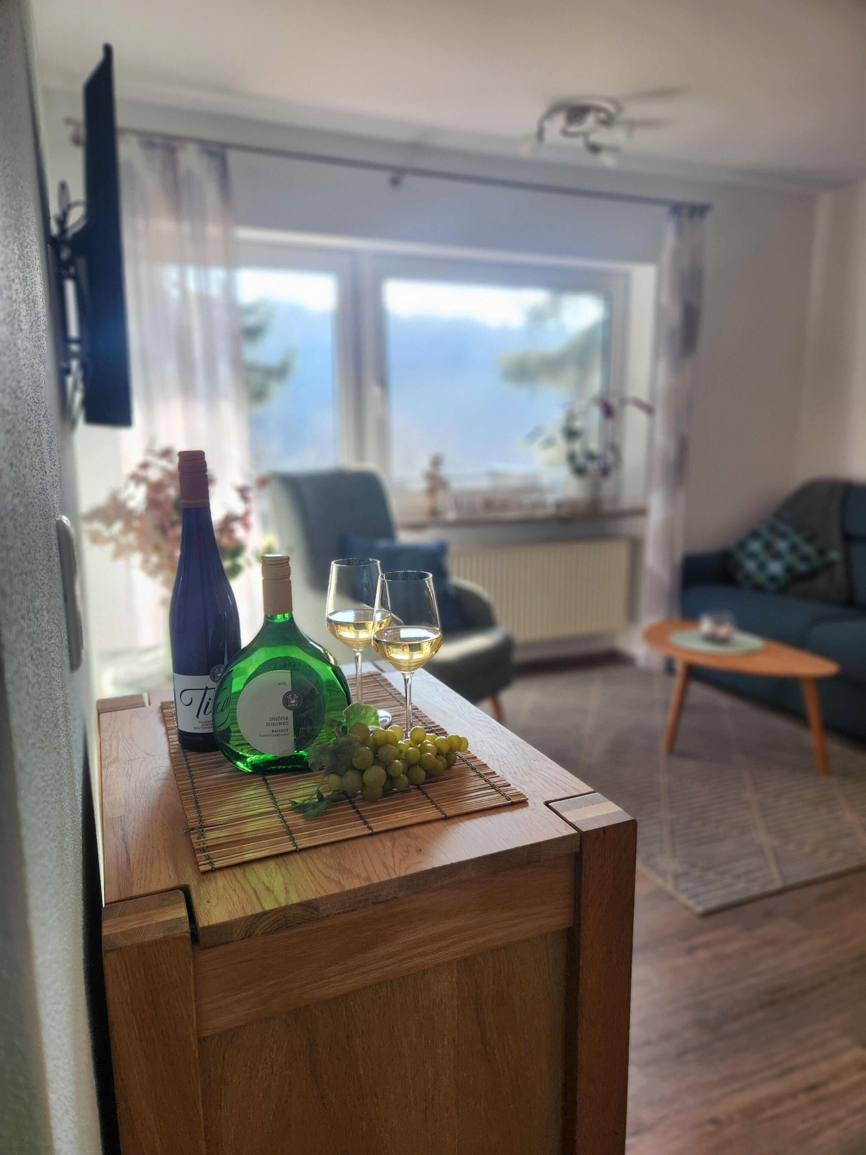 Ein Zimmer mit einem Holztisch, der eine Flasche, Gläser und Trauben trägt, einer Couch, einem Sessel und Meerblick durch ein Fenster.