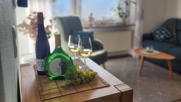 Ein Zimmer mit einem Holztisch, der eine Flasche, Gläser und Trauben trägt, einer Couch, einem Sessel und Meerblick durch ein Fenster.