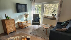 A cozy living room with a sofa, a wooden side table, a TV mounted on the wall, and a window with a view of trees. - Holiday Apartment 'Bacchus' with Private Balcony, Shared Terrace (Iphofen)