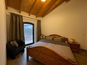 Ein Schlafzimmer mit einem hölzernen Bett, einem kleinen Nachttisch, einem Sessel und einem Fenster mit Vorhängen.
