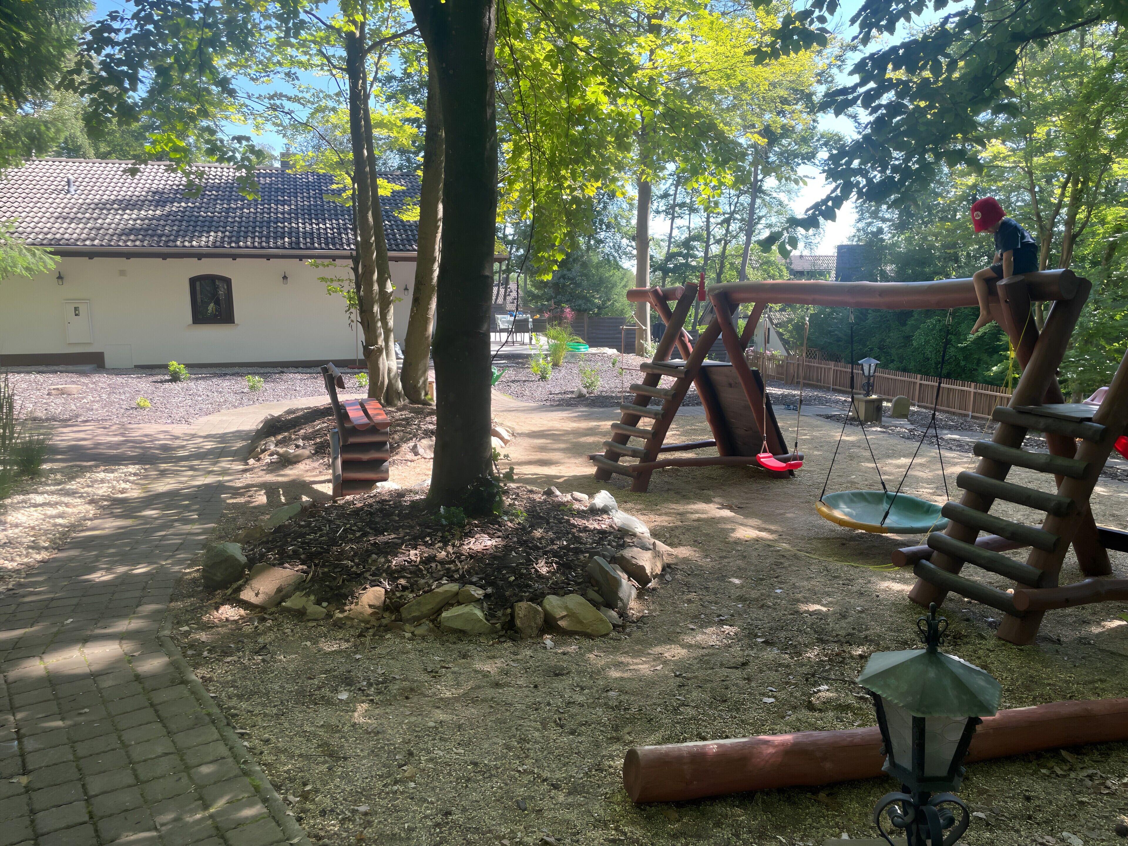 Ein Spielplatz mit Schaukeln und Rutsche, umgeben von Bäumen und einem Gebäude mit Ziegeldach.