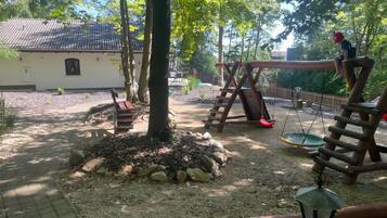 Ein Spielplatz mit Schaukeln und Rutsche, umgeben von Bäumen und einem Gebäude mit Ziegeldach.