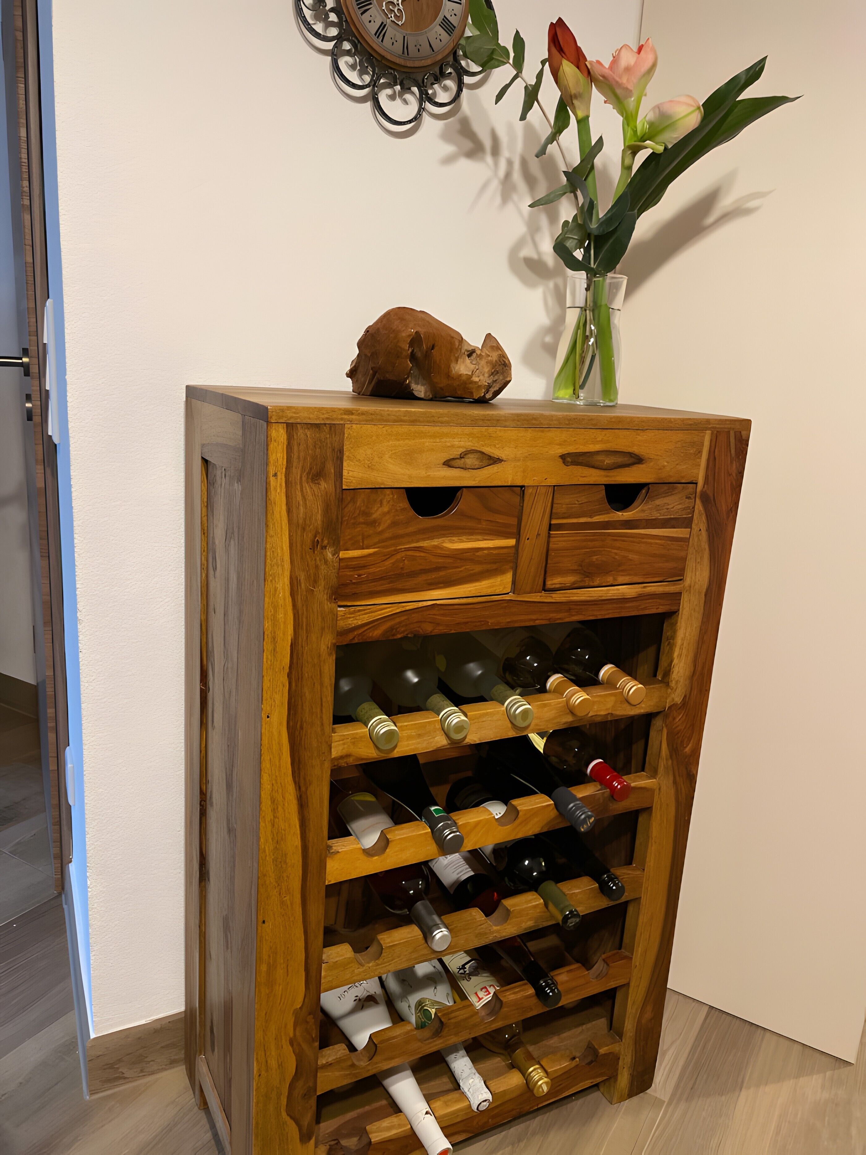 Een houten wijnrek met meerdere planken, een decoratief voorwerp bovenop, en een vaas met bloemen op de achtergrond.