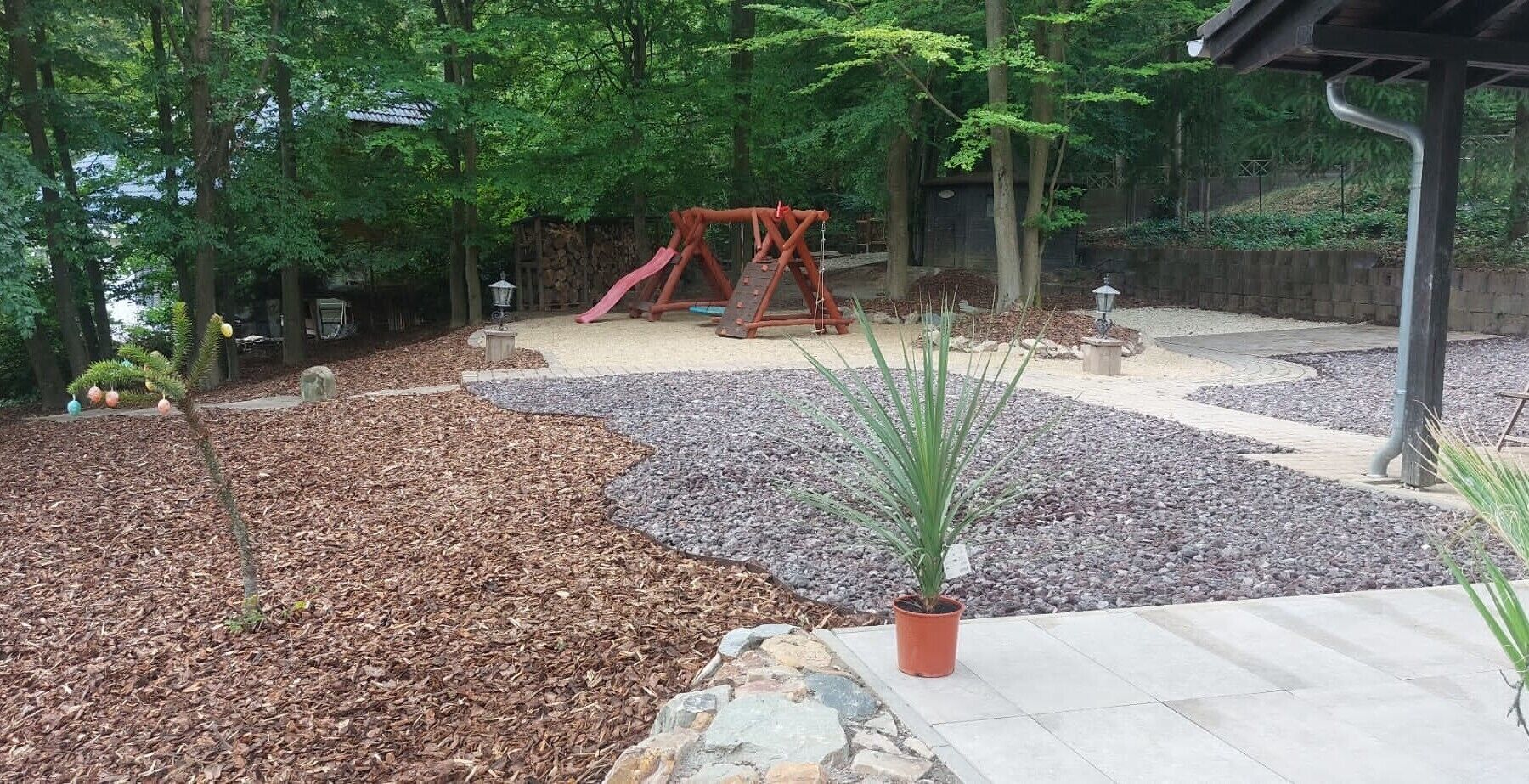 Een speelplaats met een schommel en glijbaan, omringd door bomen en een mulchgrond.