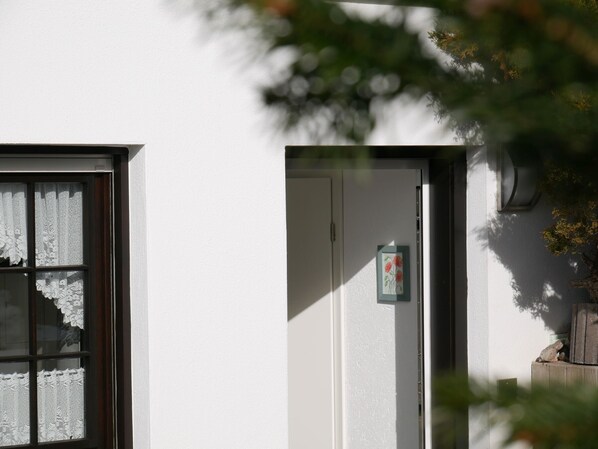 Eine weiße Außenwand mit einem Fenster, das mit Spitzenvorhängen versehen ist, und einer Tür mit einer kleinen Blumendekoration.