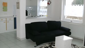 Una sala de estar moderna con un sofá negro, una mesa de centro blanca, una zona de cocina con armarios blancos y un ventanal con vistas a árboles.