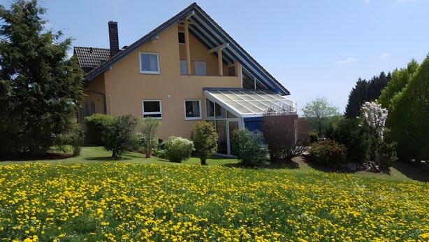 Una casa de dos pisos con techo inclinado, un invernadero y un jardín con flores amarillas.