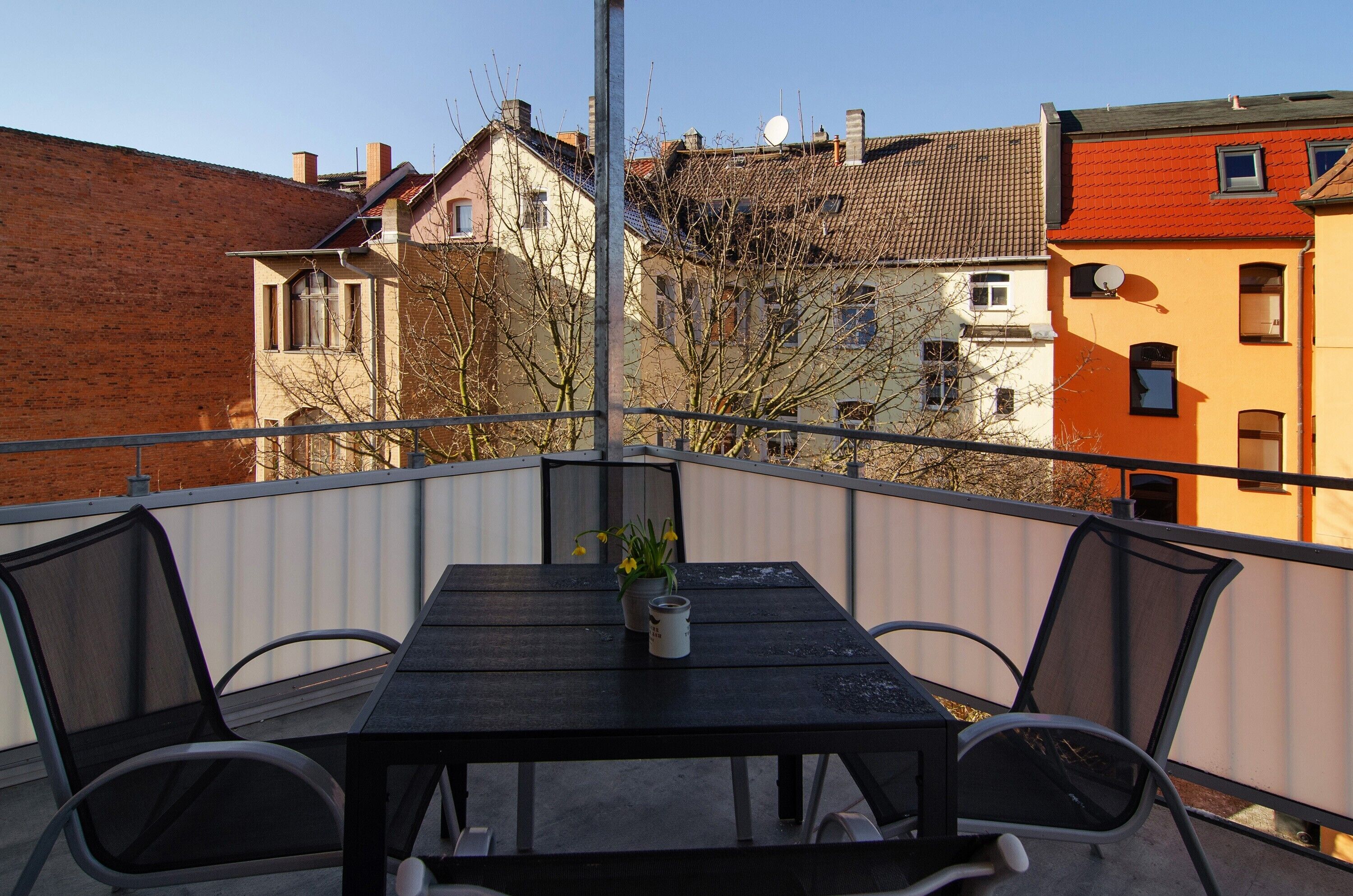 A balcony with a table and chairs, overlooking a row of buildings with varied architecture.
