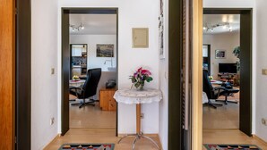 A hallway with a round table with a lace tablecloth, a mirror, and a door leading to a room with a desk, chair, and a television. - Roodbach Apartment: Mountain View, Private Terrace, Garden, Table Tennis & Wi-Fi (Lenzkirch)