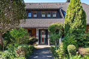 Ein zweistöckiges Haus mit Ziegeldach, einer Haustür mit roter Streifenverzierung und einem Garten mit verschiedenen Pflanzen und einem hohen Baum.