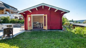 A red wooden building with an open door, a grill, and a bench outside. - Roodbach Apartment: Mountain View, Private Terrace, Garden, Table Tennis & Wi-Fi (Lenzkirch)