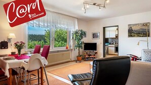 A modern living room with a dining area, a TV, and a view of greenery outside. - Roodbach Apartment: Mountain View, Private Terrace, Garden, Table Tennis & Wi-Fi (Lenzkirch)