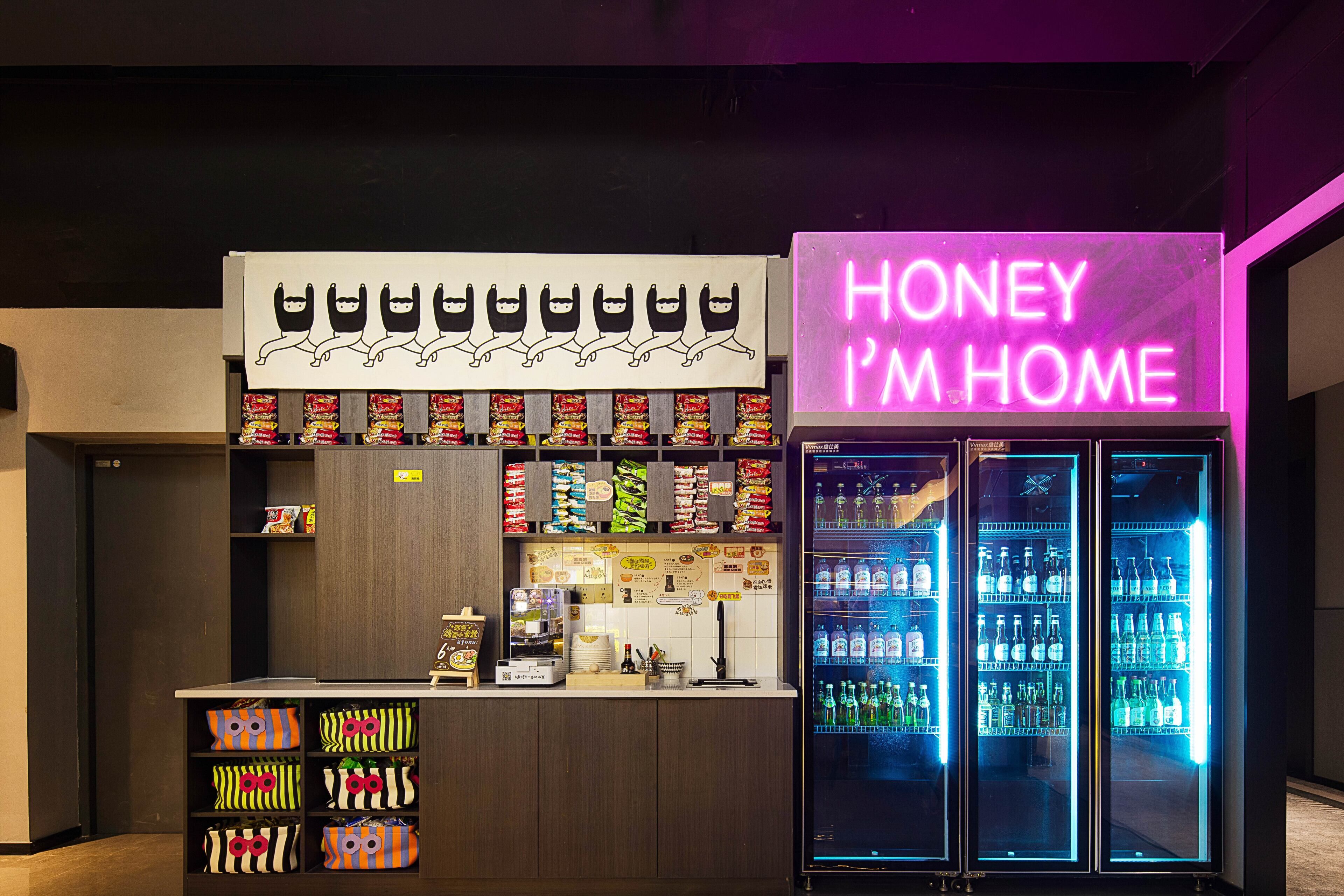 A bar area with a neon sign reading "HONEY I'M HOME," shelves stocked with snacks, and a refrigerator filled with beverages.