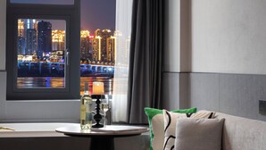 A modern hotel room with a view of a city skyline at night, a round table with a candle, and a sofa with decorative pillows.