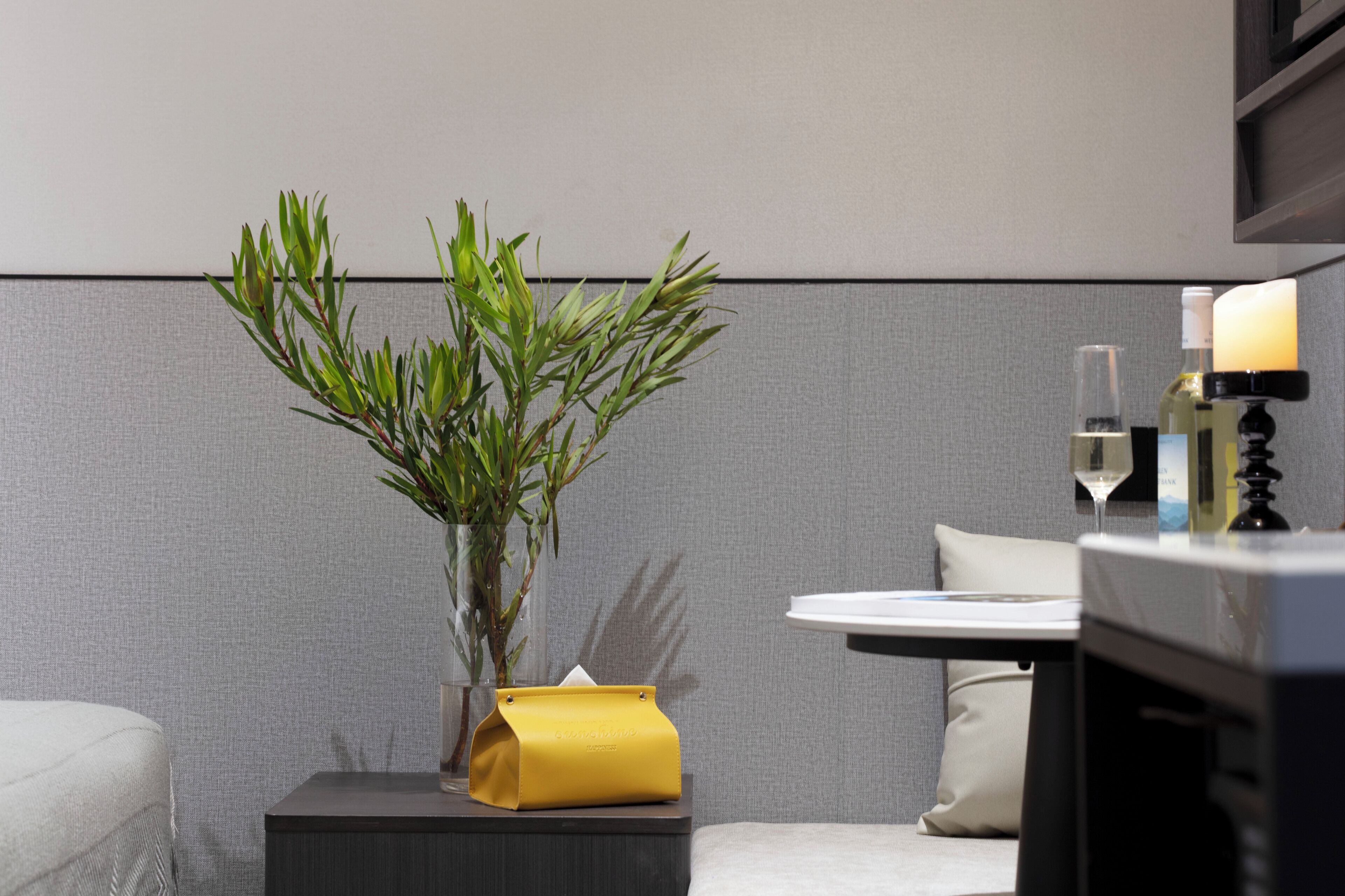 A modern room with a grey textured wall, a small bedside table with a yellow tissue box, a bedside plant, a white pillow, and a bedside lamp.