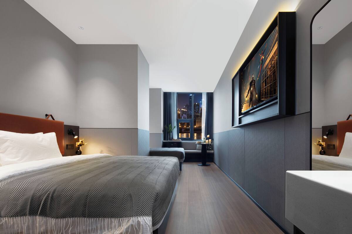 A modern hotel room with a large bed, a desk, and a view of the cityscape through a window.