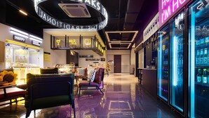 A modern hotel lobby with a neon sign reading "HONEY I'M HOME", a seating area with a coffee machine, and a refrigerator stocked with beverages.