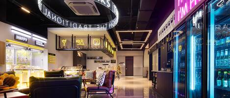 A modern hotel lobby with a neon sign reading "HONEY I'M HOME", a seating area with a coffee machine, and a refrigerator stocked with beverages.