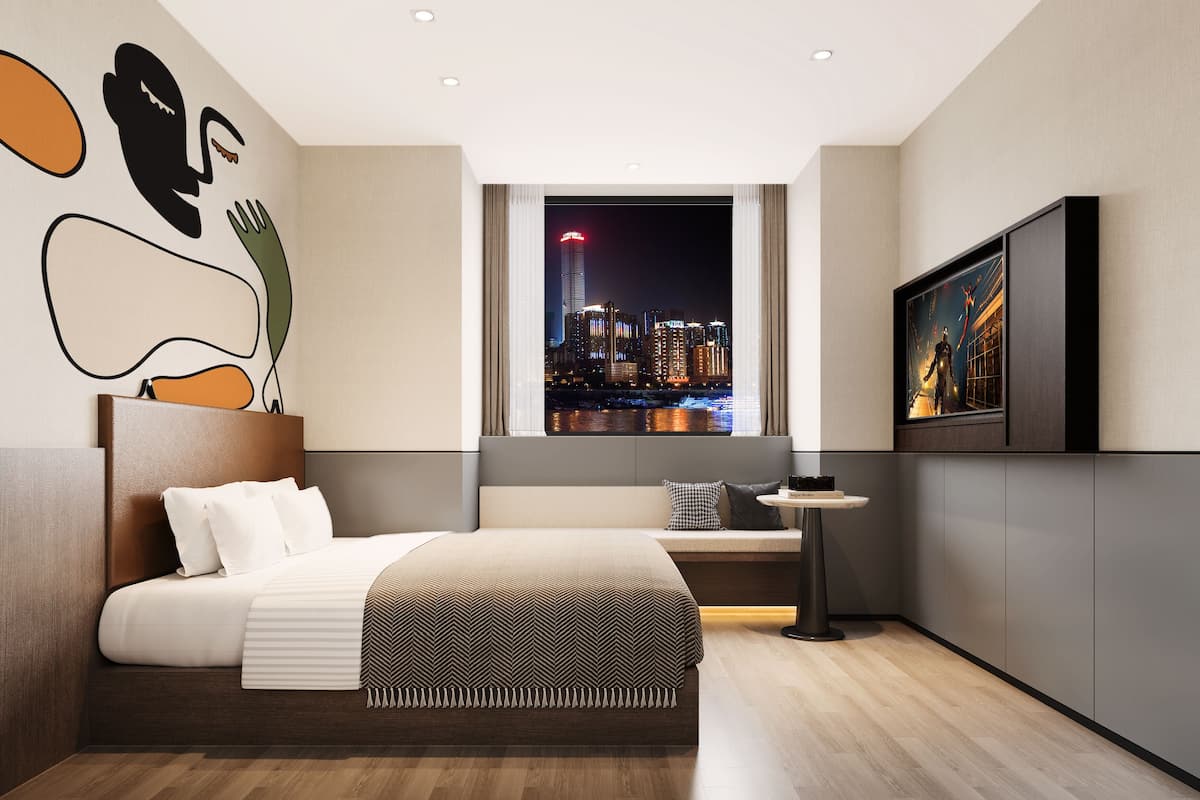 A modern hotel room with a large bed, a view of a city skyline at night, a flat-screen TV, and a wall with abstract art.