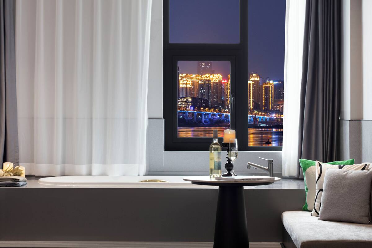 A modern hotel room with a large window offering a nighttime city view, a bathtub, and a small table with a bottle and glass.