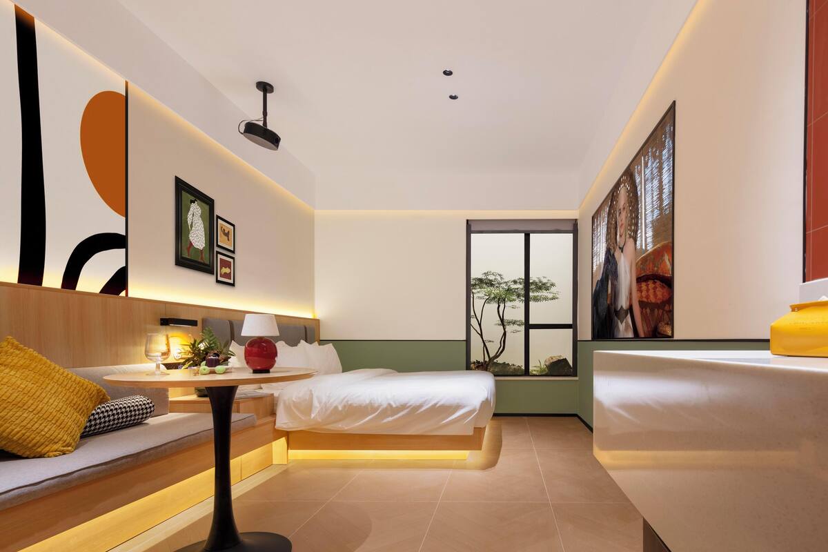 A modern hotel room with a bed, a sofa, a table with a lamp, and artwork on the walls.