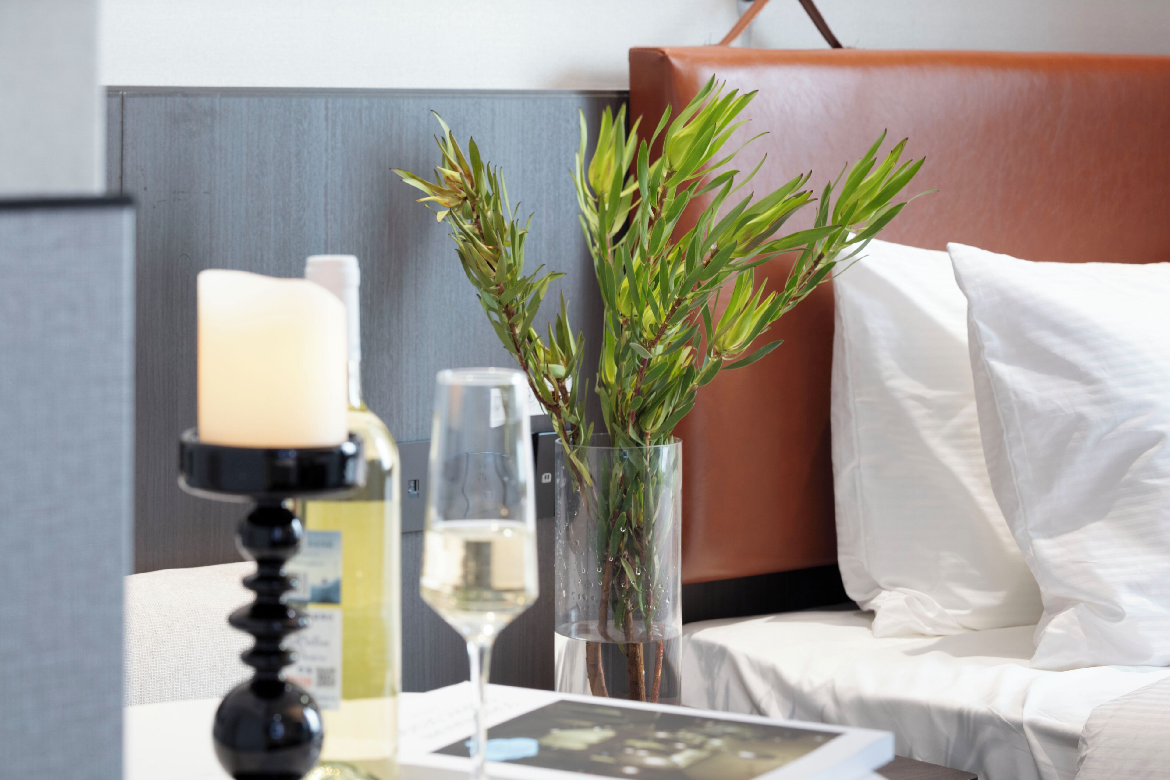 A neatly arranged hotel room with a bedside table featuring a candle, a glass of wine, and a vase with greenery.