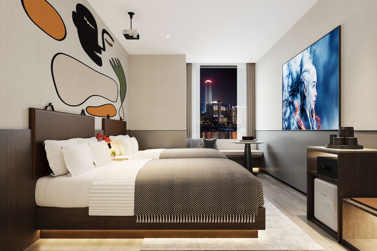 A modern hotel room with a large bed, a city view, a projector, and a large artwork on the wall.