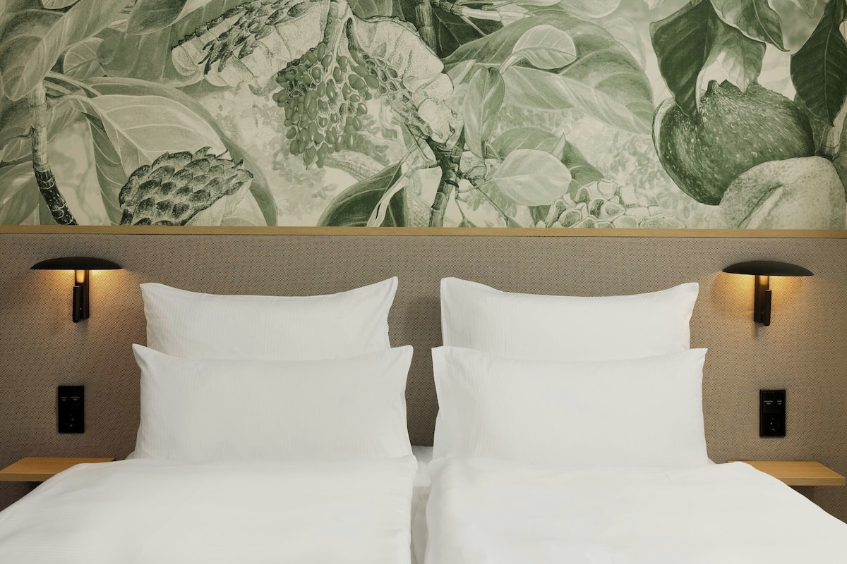 A hotel room with a bed, two bedside tables, and a wall with a botanical mural.