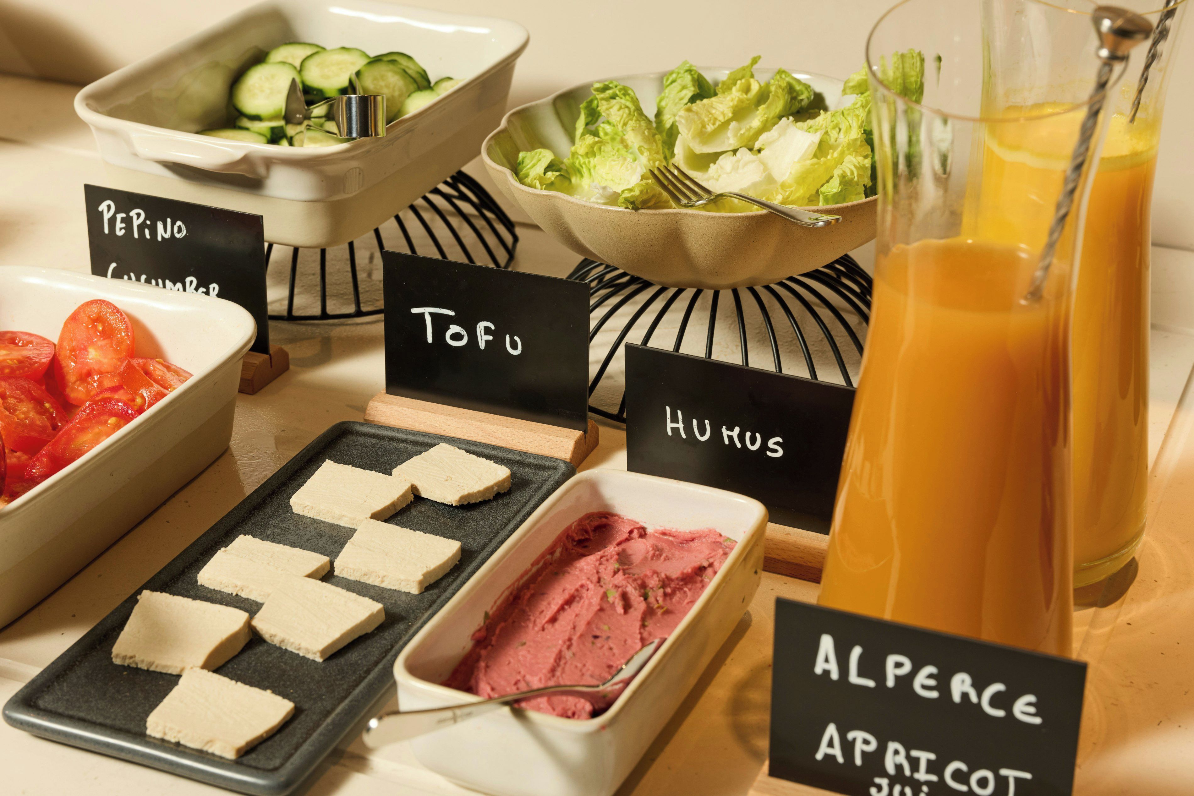 Un buffet proposant une variété de plats, chacun étant clairement identifié, comprenant des concombres, de la laitue, des tomates, du tofu, du houmous et une boisson.
