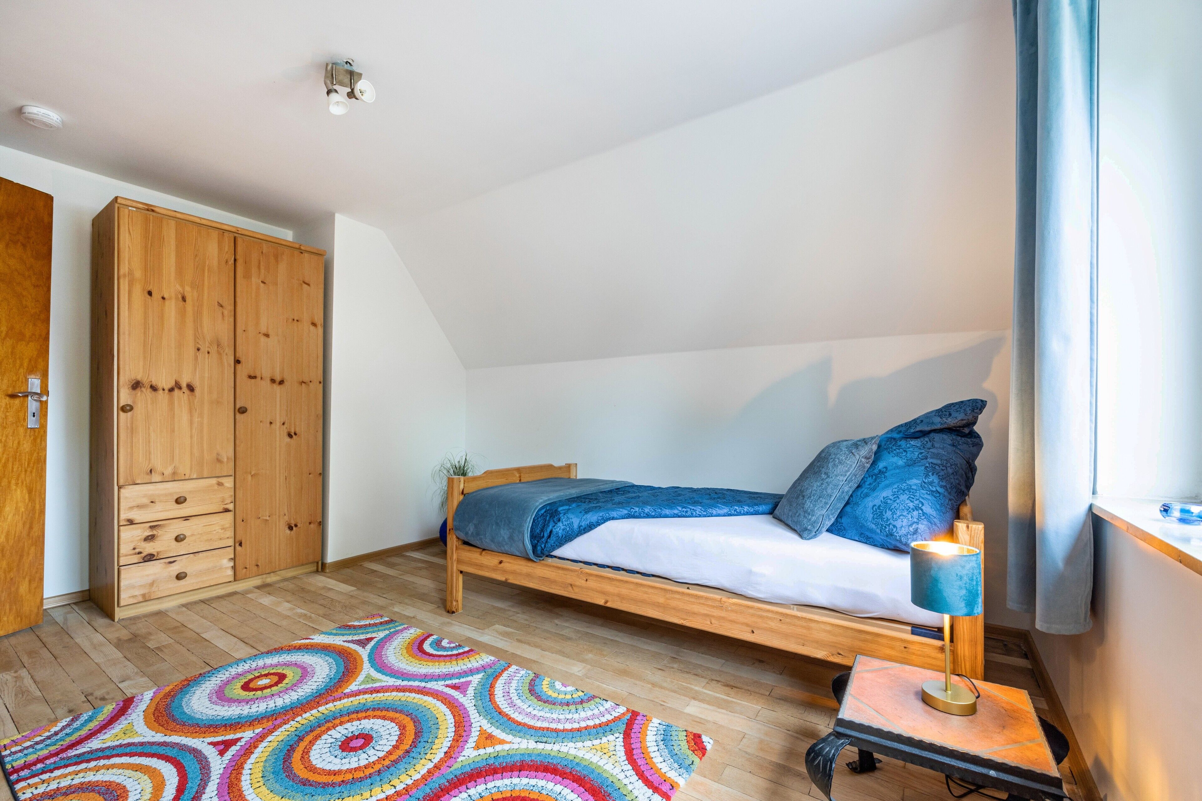 Un dormitorio con una cama de madera, una alfombra colorida, una mesita con una lámpara y un armario.