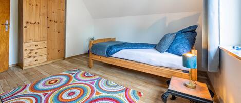 Un dormitorio con una cama de madera, una alfombra colorida, una mesita con una lámpara y un armario.
