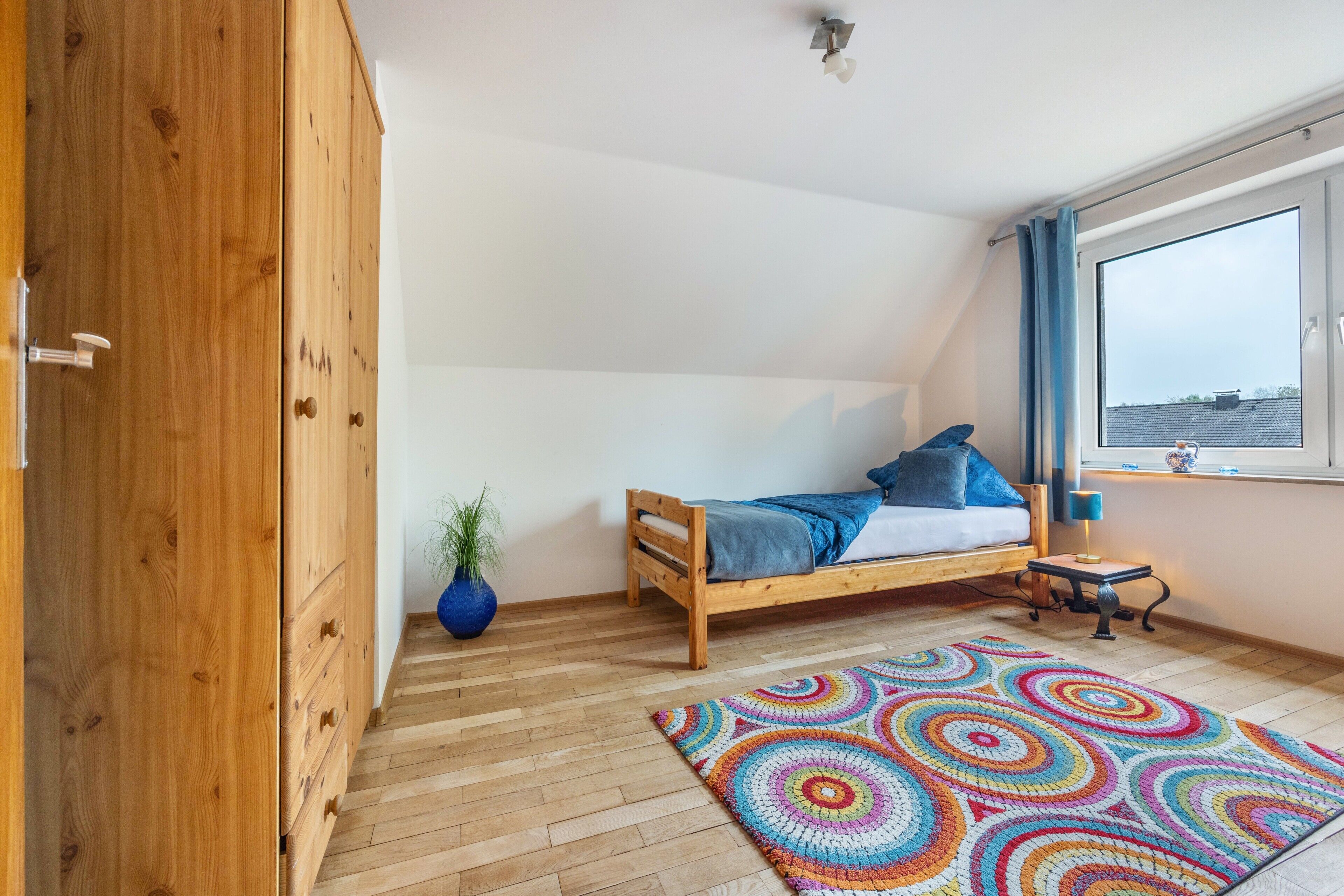 A bedroom with a wooden bed, a colorful rug, a potted plant, and a window with curtains.