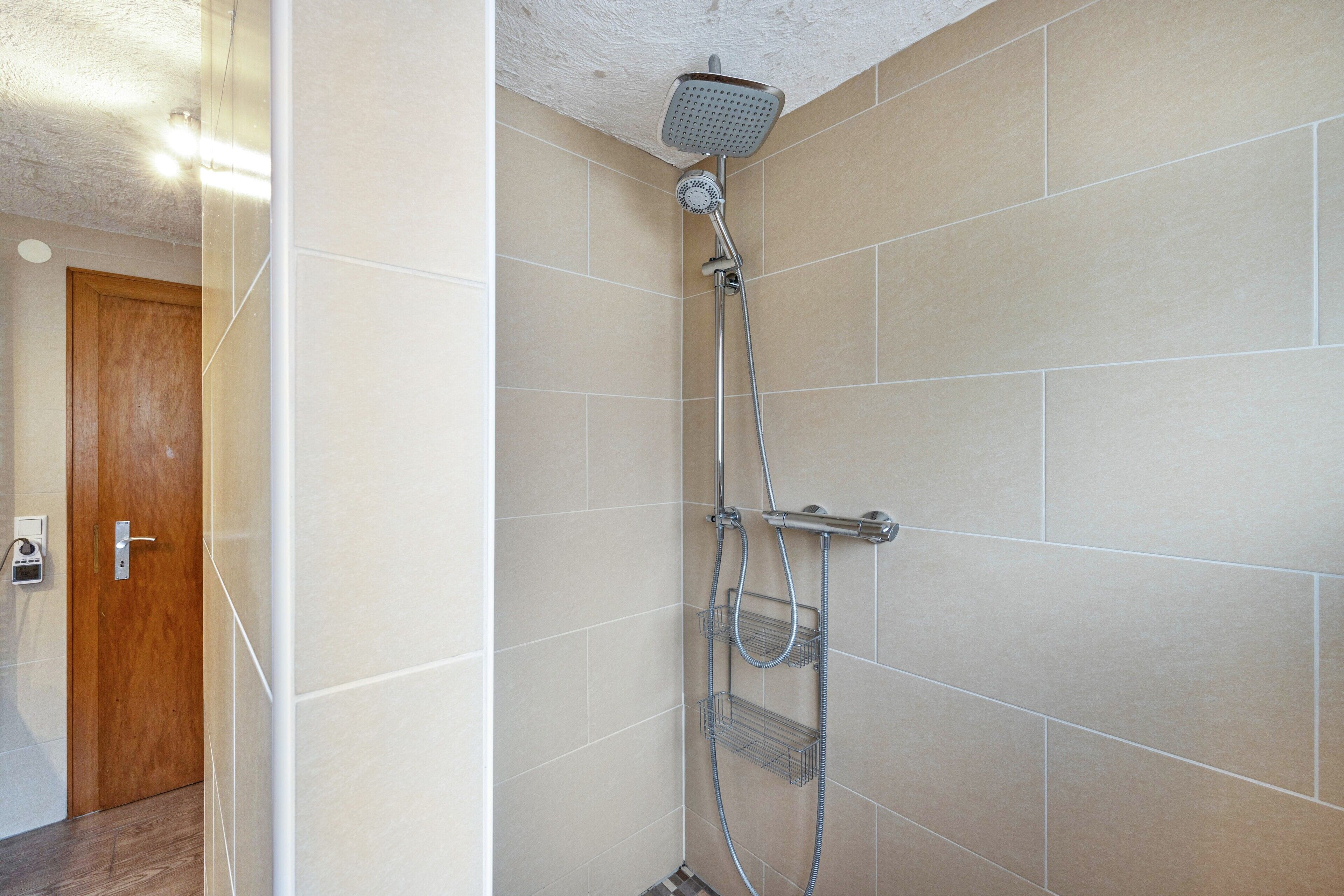 Une douche équipée d’un pommeau de douche à main et d’un robinet mural, une porte avec une poignée et un mur recouvert de carreaux.