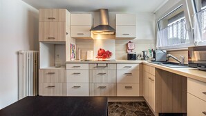 A modern kitchen with light wood cabinets, a central range hood, a black countertop, and a window with a view.