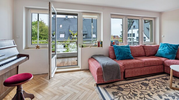 A room with a piano, a red sofa with blue cushions, a patterned rug, and large windows with a view of buildings and trees.