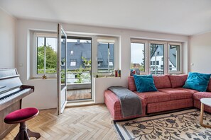 Una habitación con un piano, un sofá rojo con cojines azules, un tapete estampado y grandes ventanas con vistas a edificios y árboles.