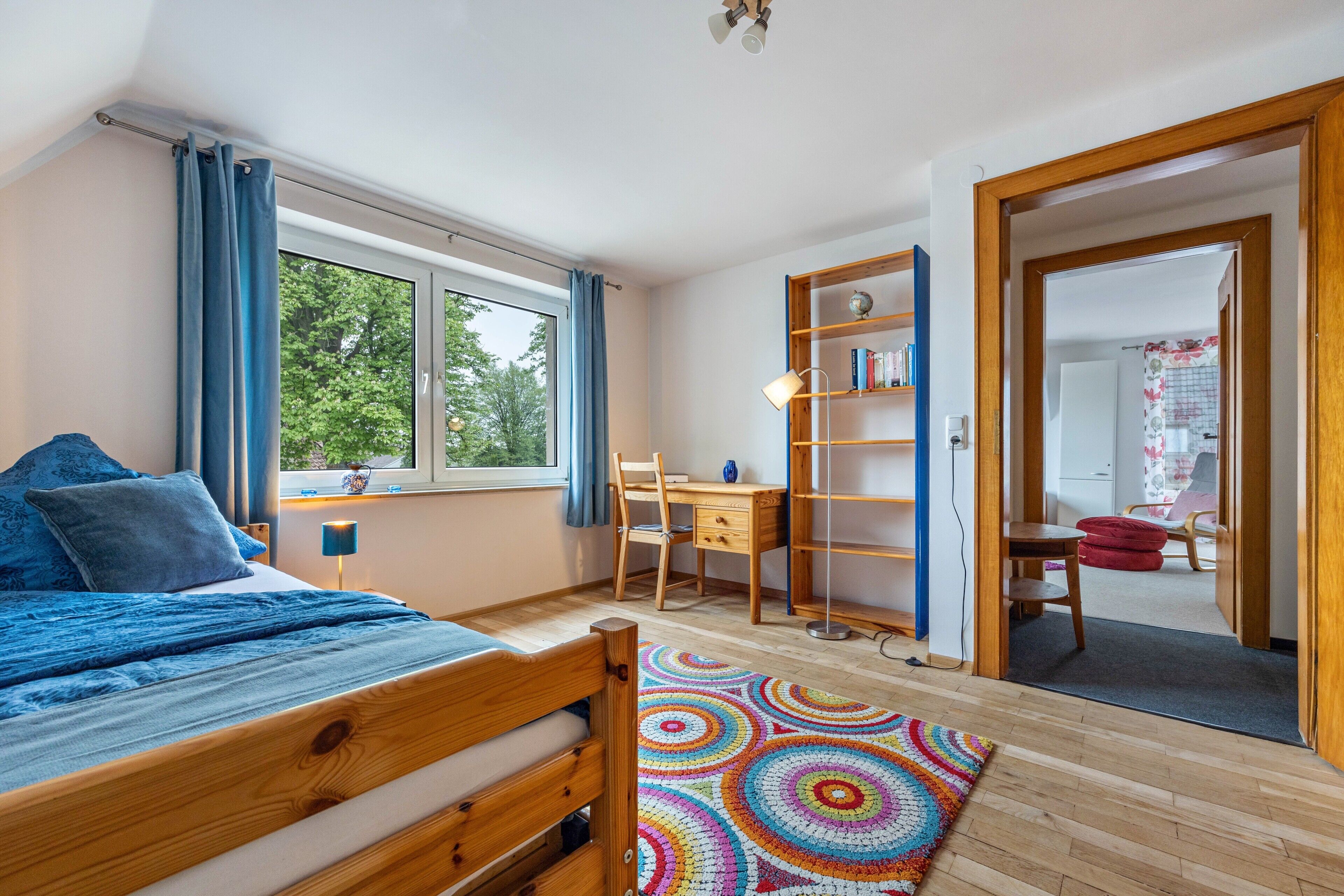A bedroom with a bed, desk, bookshelf, and a view of the outdoors.