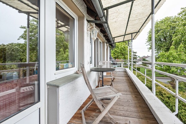 Una terraza de madera con mesa y sillas, barandilla blanca y vistas a zonas verdes.