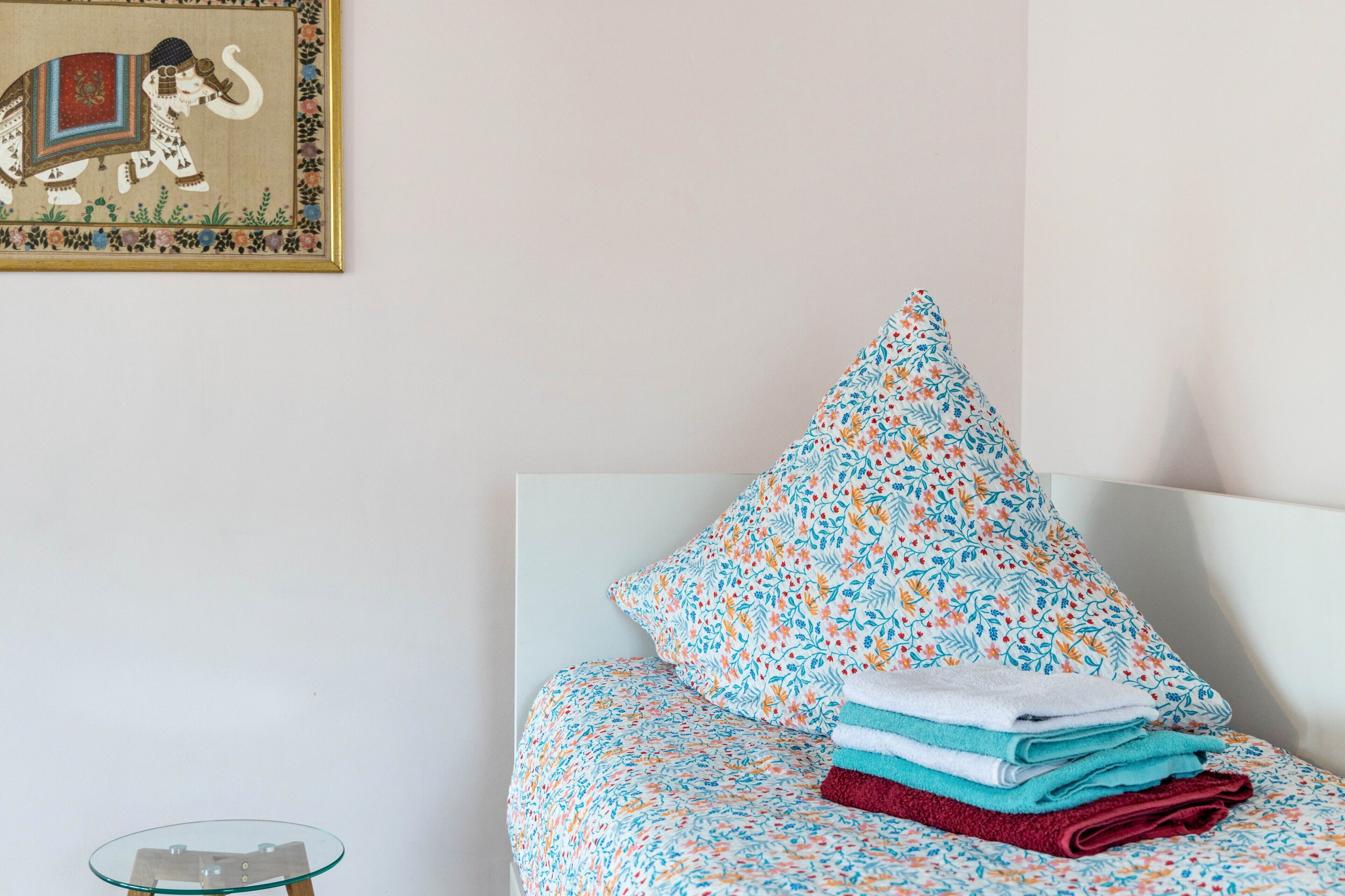 Un lit avec une literie à motifs floraux, un oreiller et des serviettes pliées. On aperçoit une table en verre avec un pied en métal.