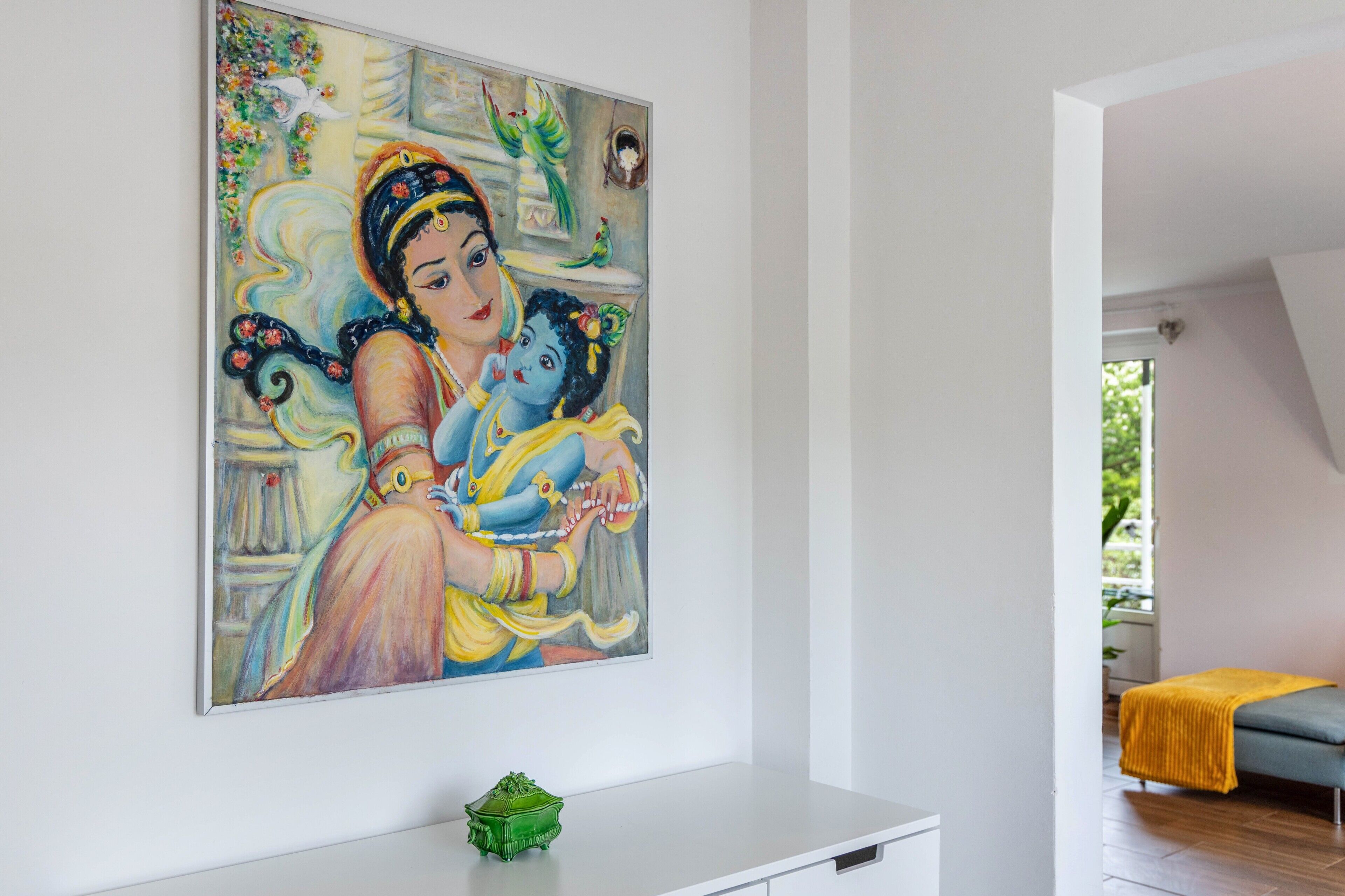 A room with a large painting, a small green object on a white cabinet, and a grey sofa with a yellow blanket.