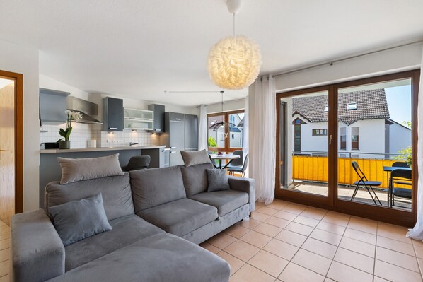 A modern living room with a grey sectional sofa, a dining area with a table and chairs, and a kitchen with grey cabinets.
