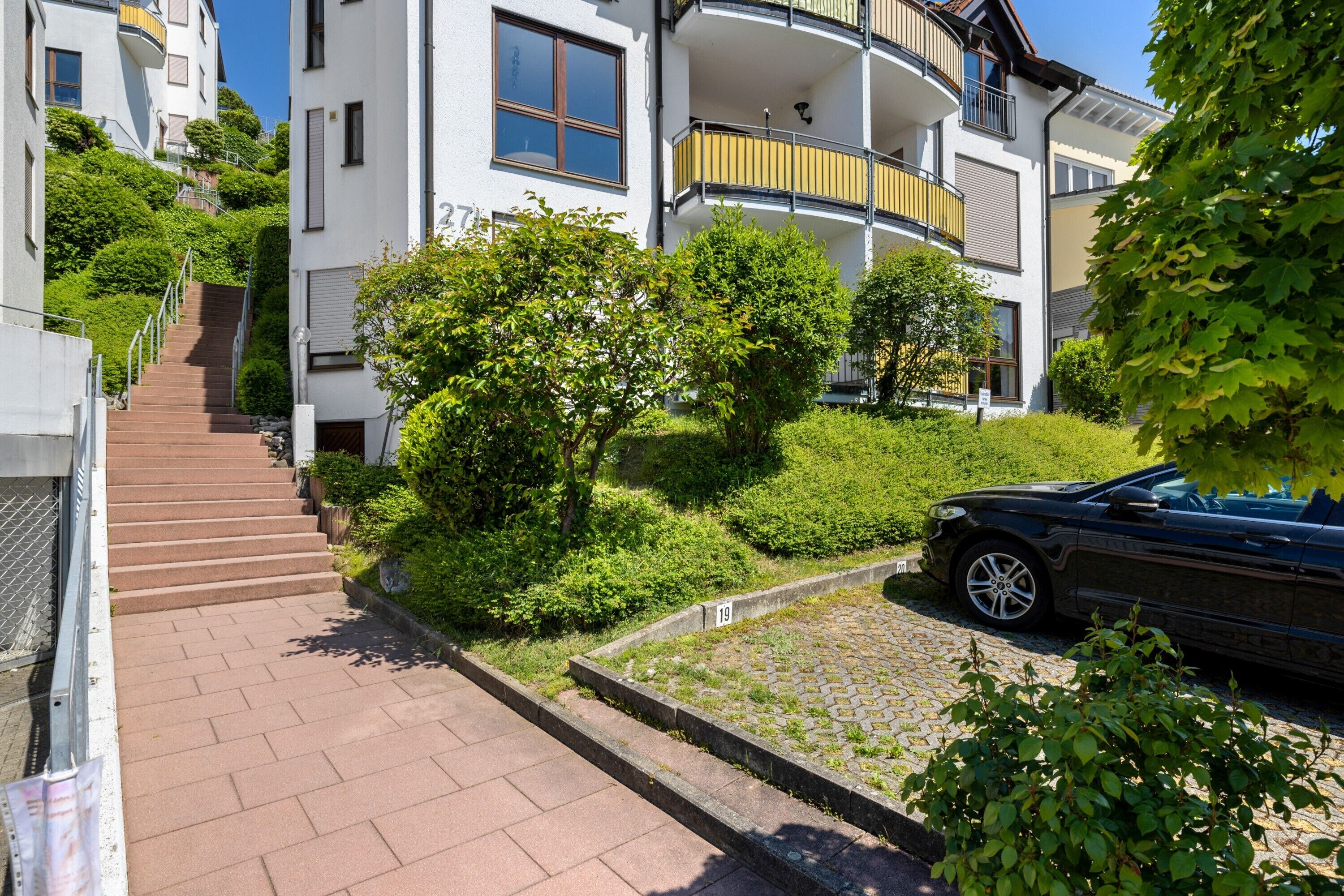 A residential building with a staircase leading to a balcony, a paved driveway, and a parked car.