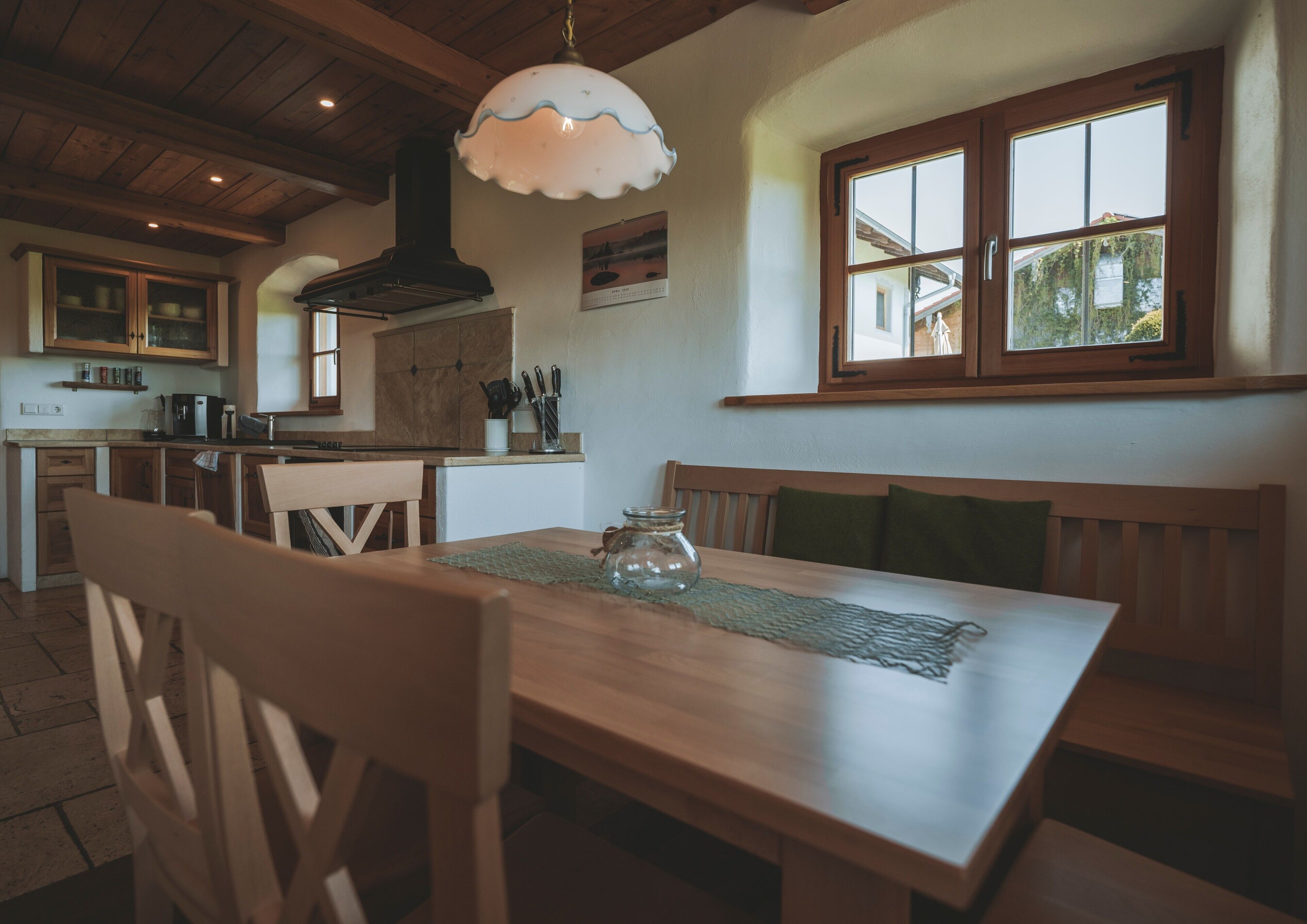 A wooden dining table with benches, a kitchen area with wooden cabinets, a hanging light fixture, and a window with a view of greenery outside.