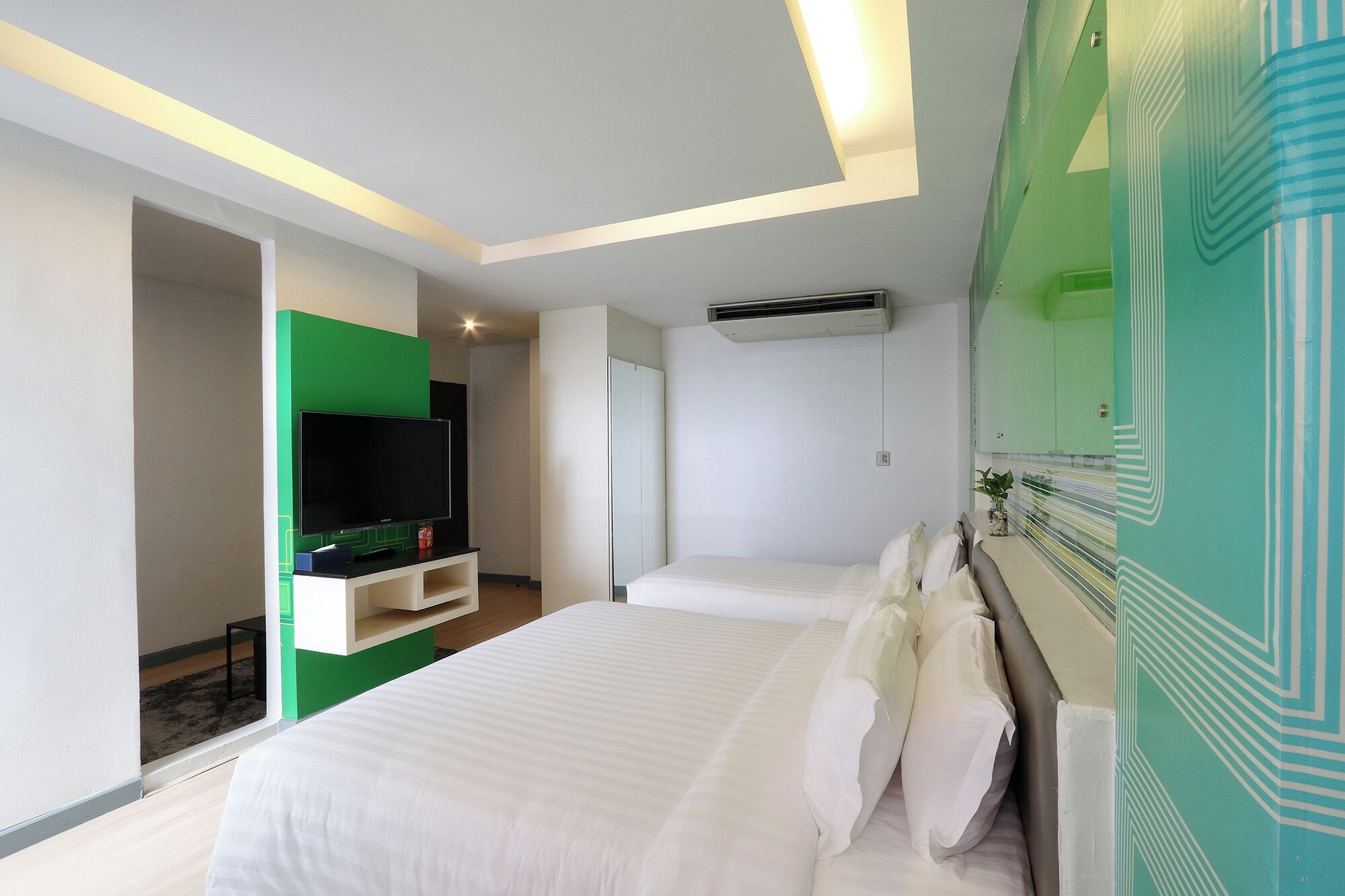 A modern hotel room with a large bed, a flat-screen TV, a green accent wall, and a ceiling-mounted air conditioning unit.