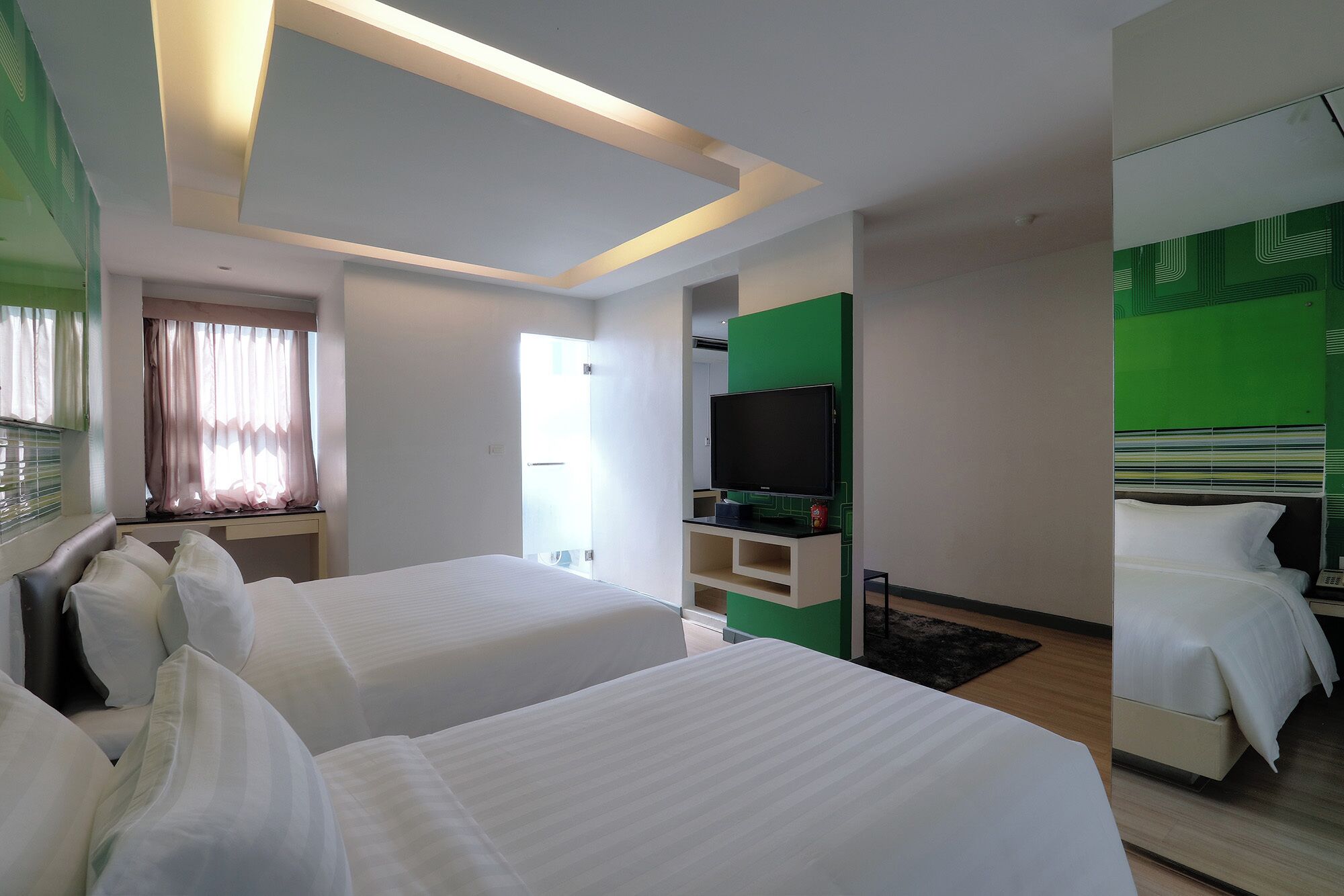 A hotel room with two beds, a flat-screen TV, a green accent wall, and a large mirror.