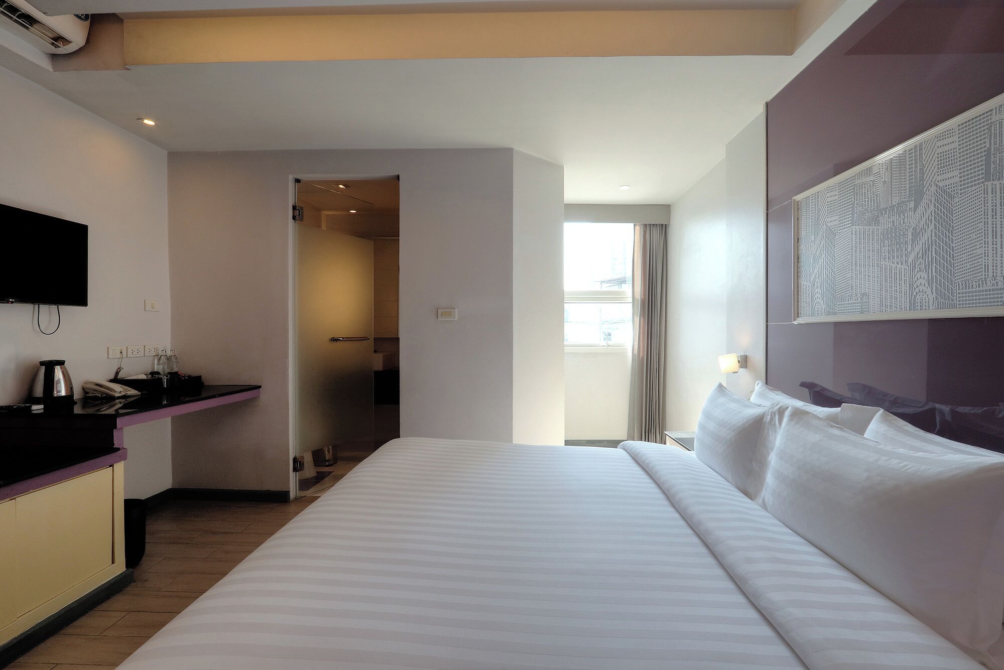 A hotel room with a large bed, a flat-screen TV mounted on the wall, a small kitchenette, and a window with curtains.