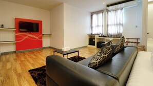 Een moderne woonkamer met een bank, een flatscreen-tv aan een rode accentmuur, een keukenhoek met een raam en een plafondventilator.