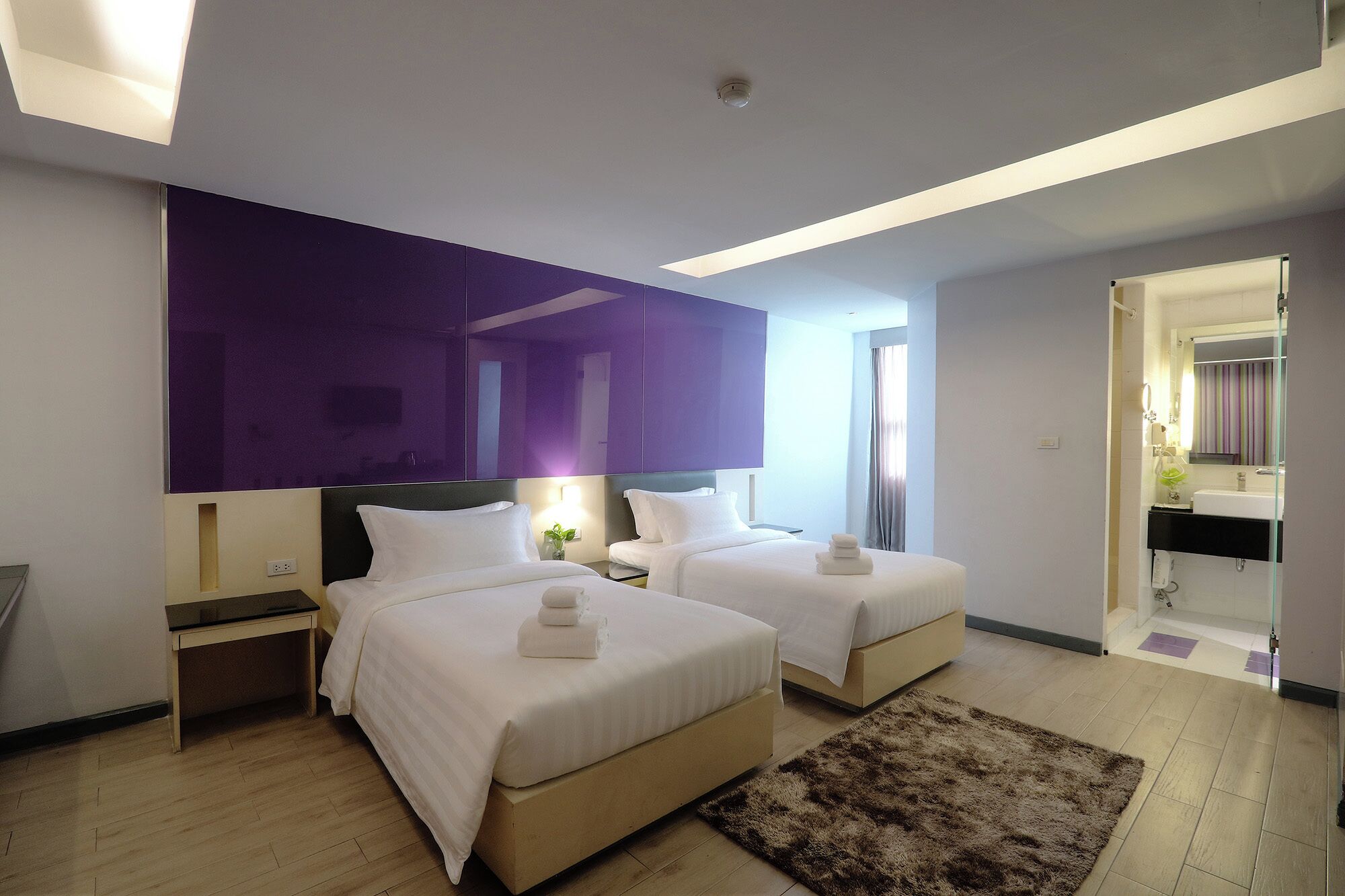 A modern hotel room with two beds, a large mirror, and a bathroom visible through an open door.