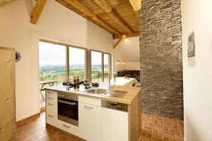 A kitchen with a large window, wooden ceiling beams, a stone accent wall, and a view of the countryside.