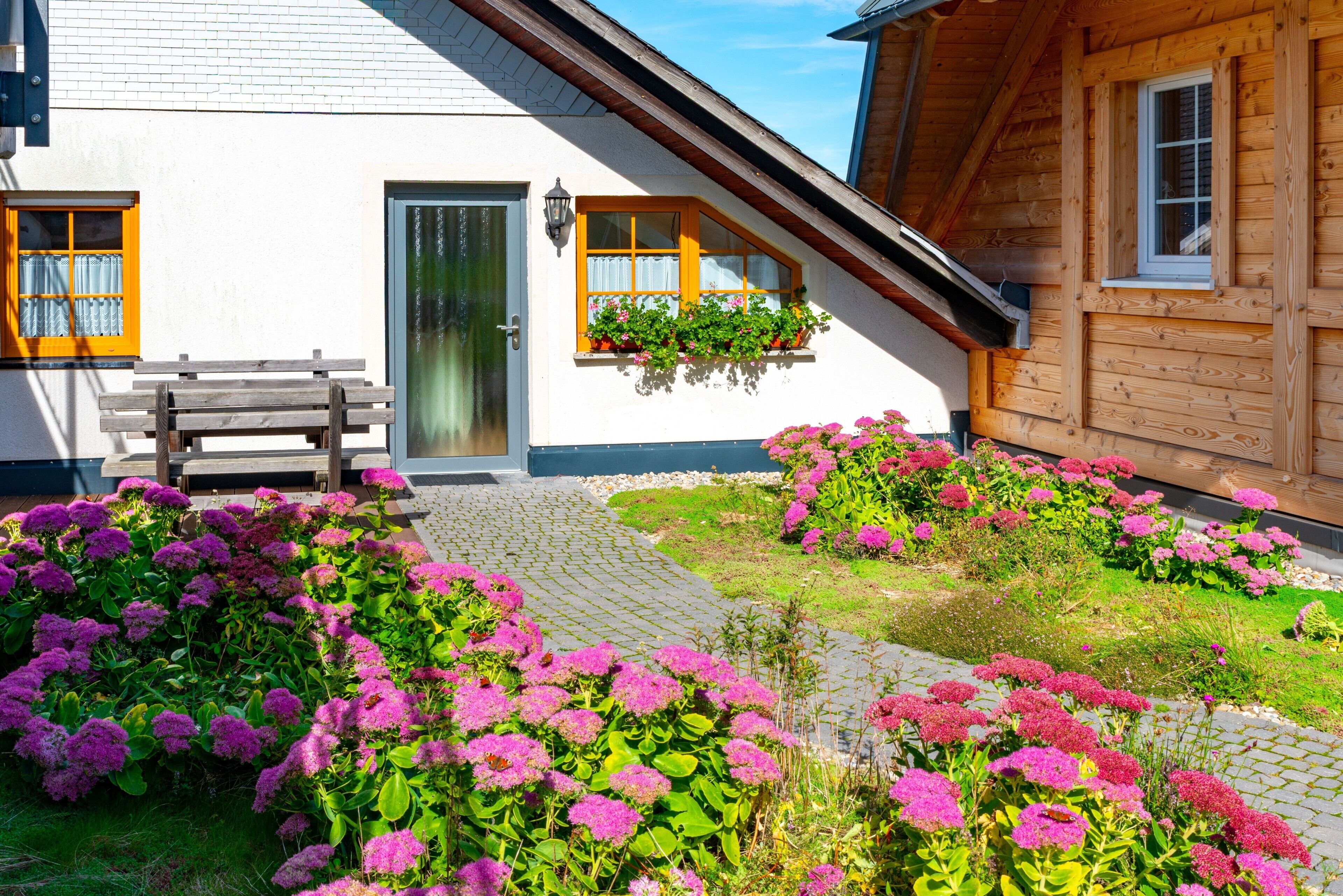 Une maison en bois à l’extérieur blanc, un banc en bois et un jardin parsemé de fleurs roses.